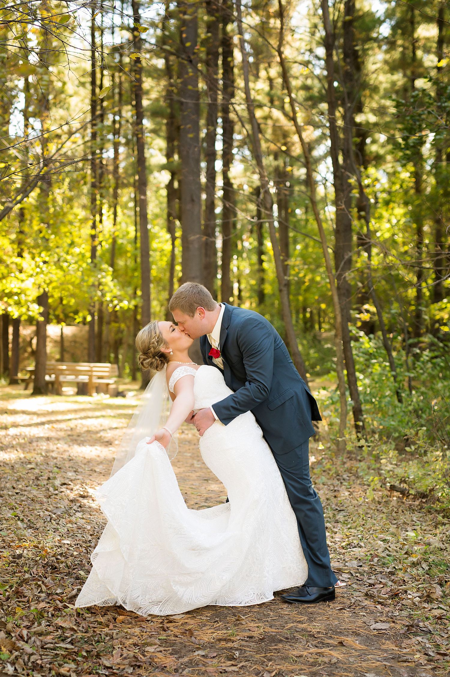 A romantic dip kiss between newlyweds in a sun-dappled forest setting.