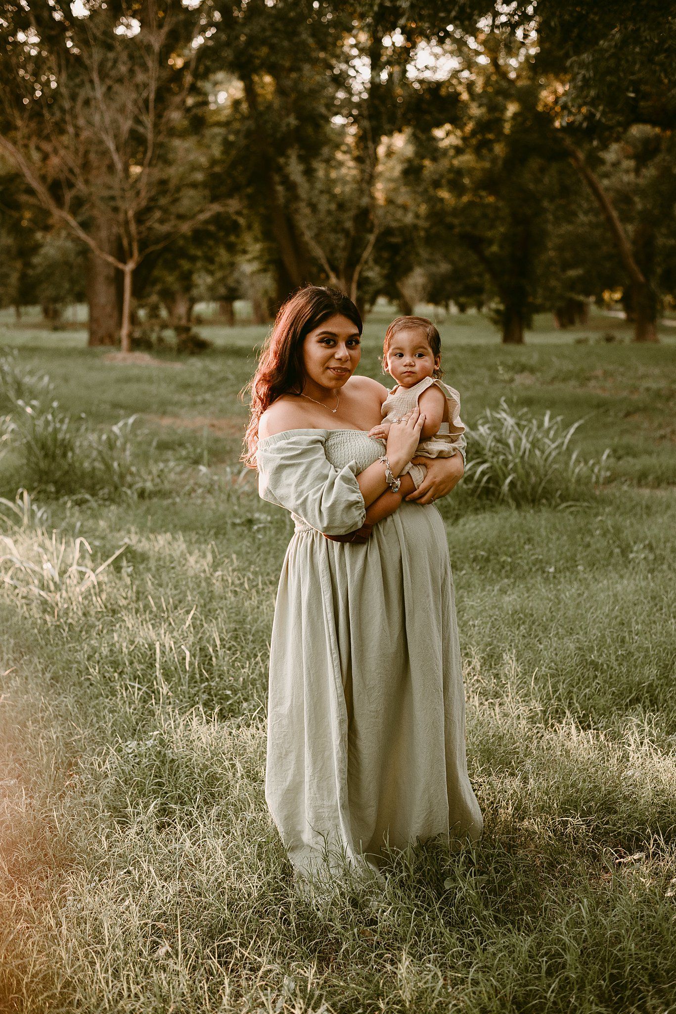 A woman in a flowing light-colored dress holds a small child in a grassy field during golden hour.
