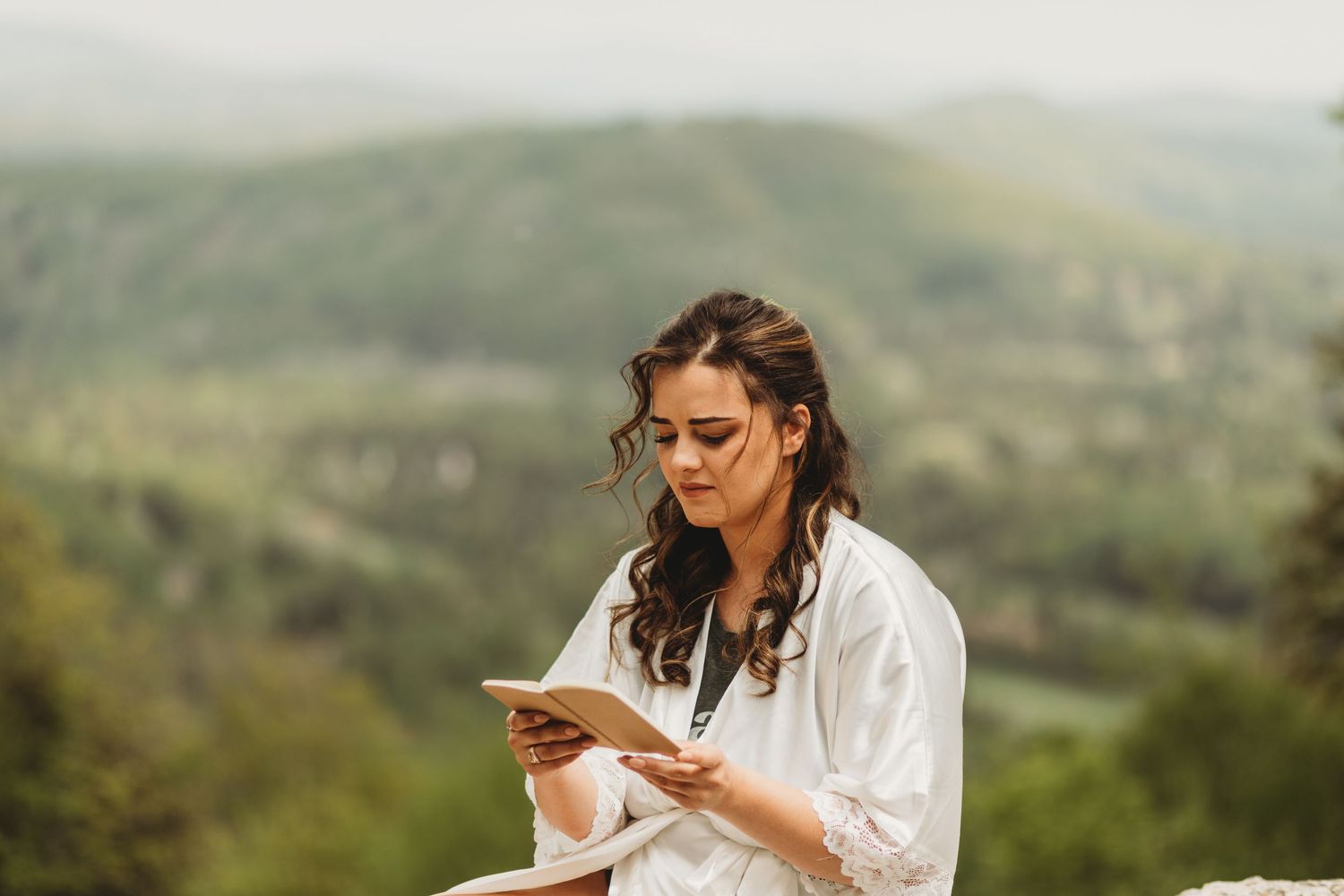 Someone in a white robe reads from a phone against a backdrop of rolling hills.