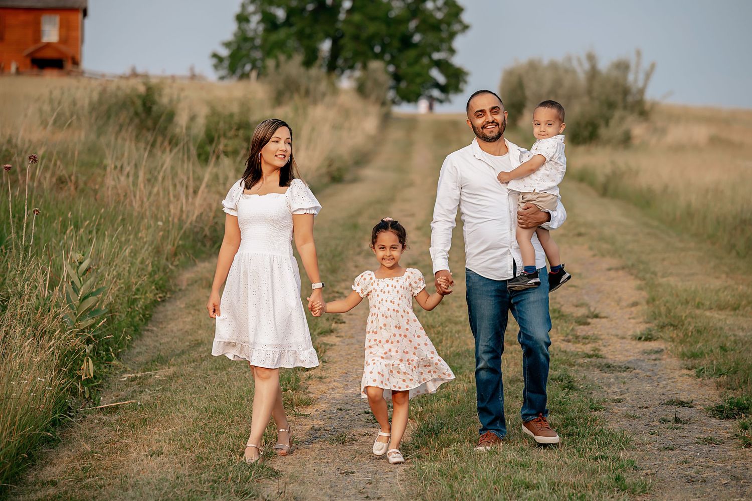 A family walks together on a dirt path through a rural summer landscape.