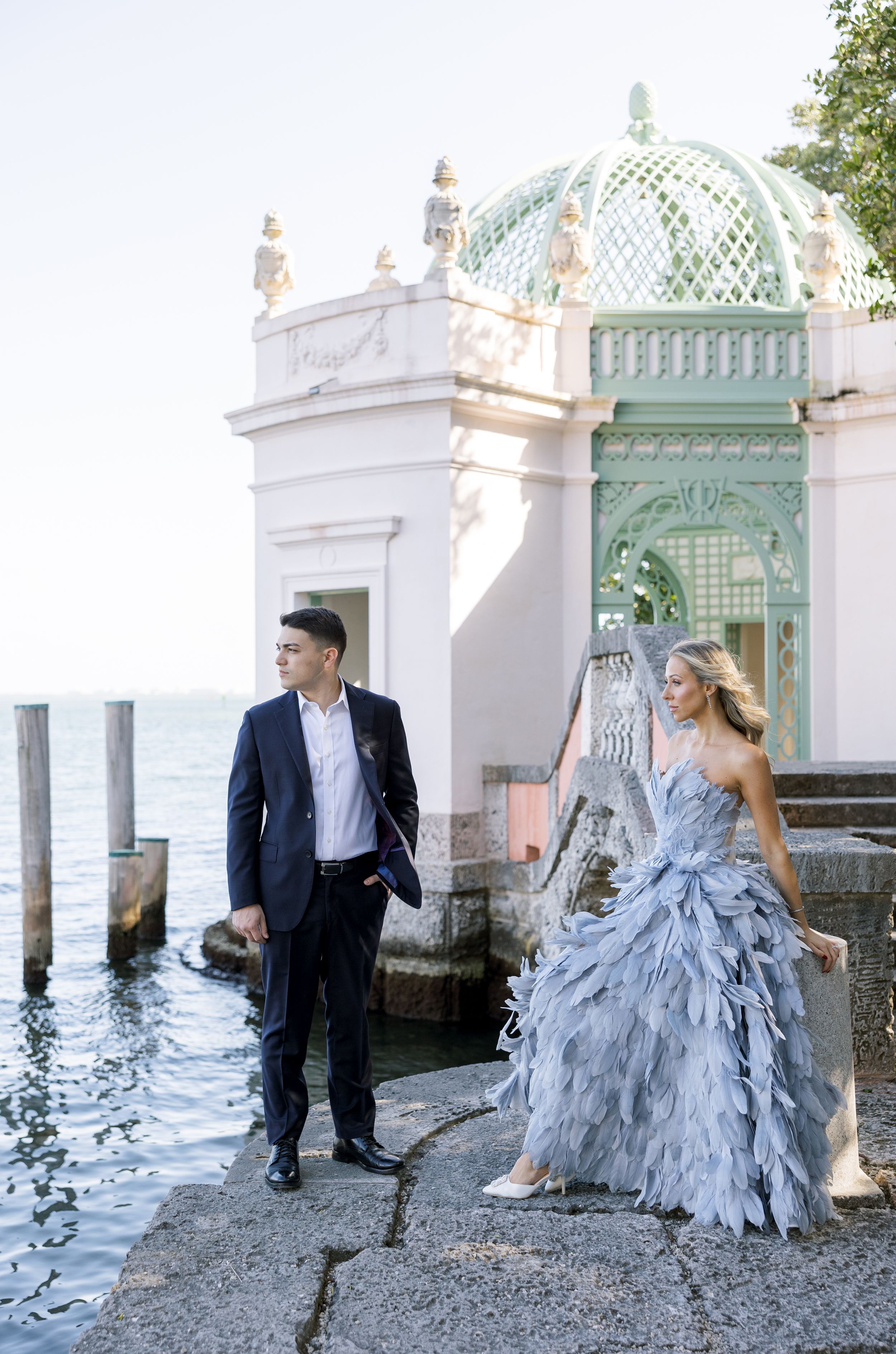A couple in elegant attire poses by a waterfront pavilion with ornate mint-green dome.