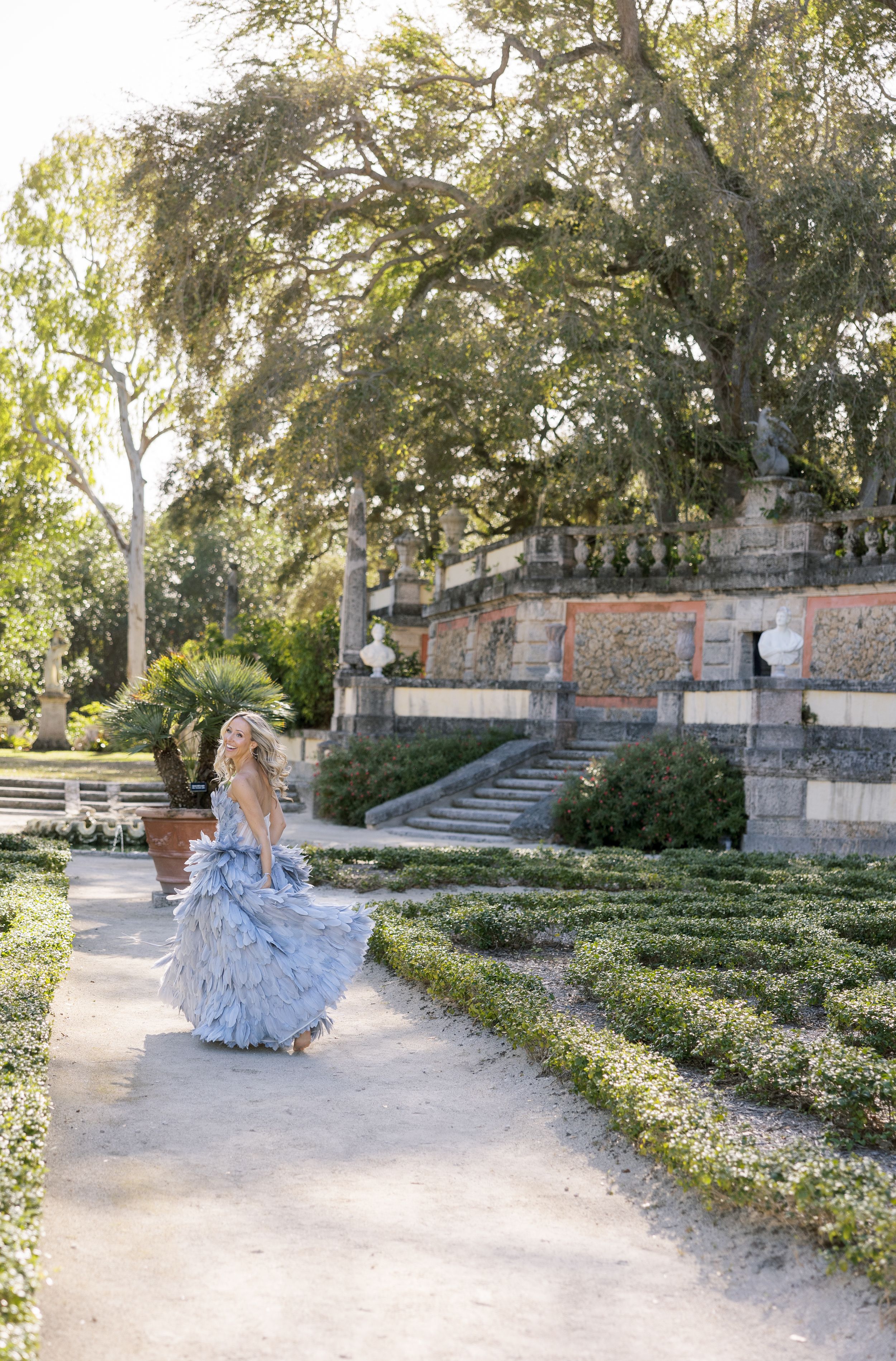 A flowing grey gown creates movement as a figure walks through a manicured garden path.