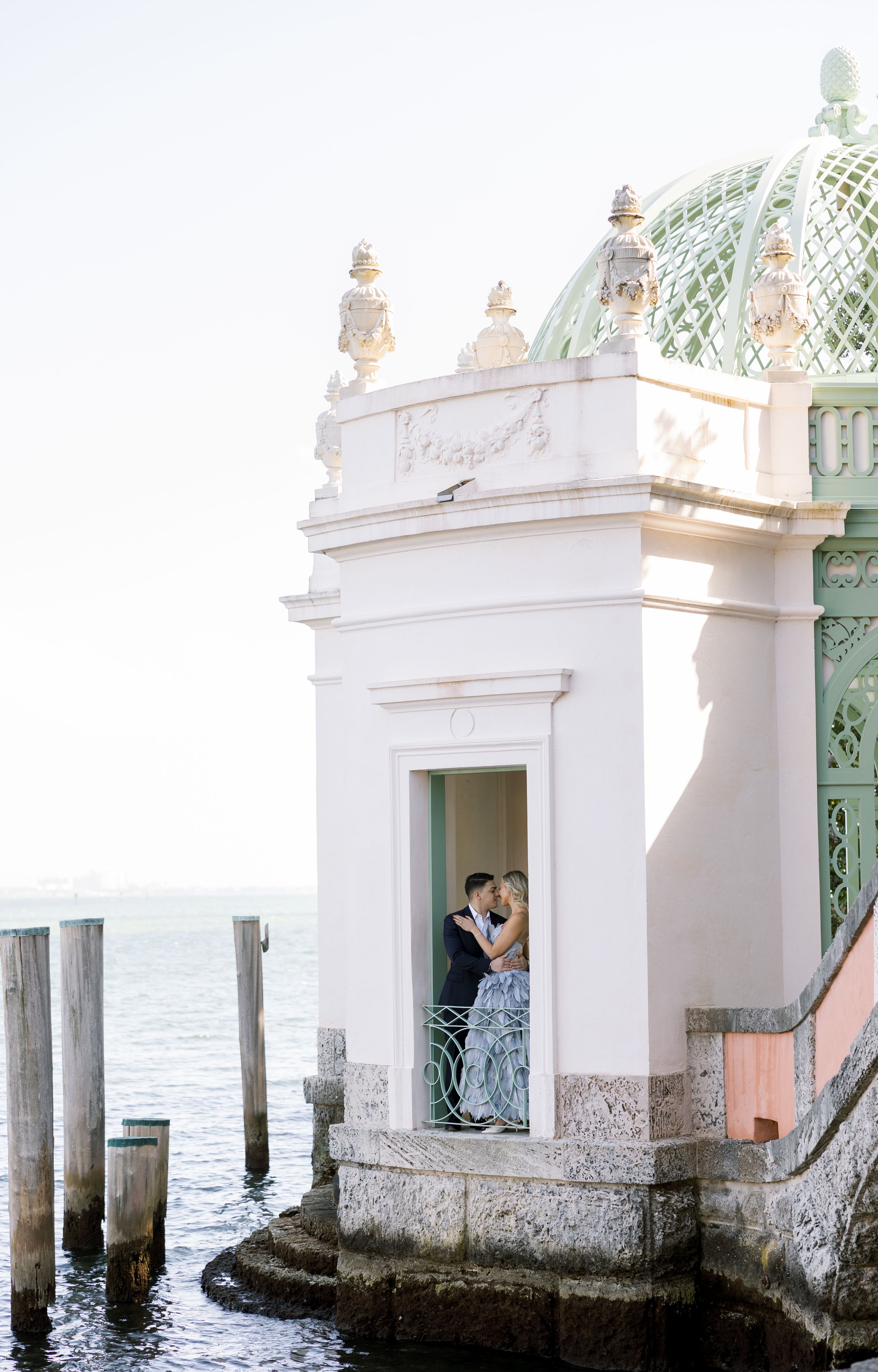 An elegant couple embraces in a waterfront gazebo with ornate architecture and turquoise details.
