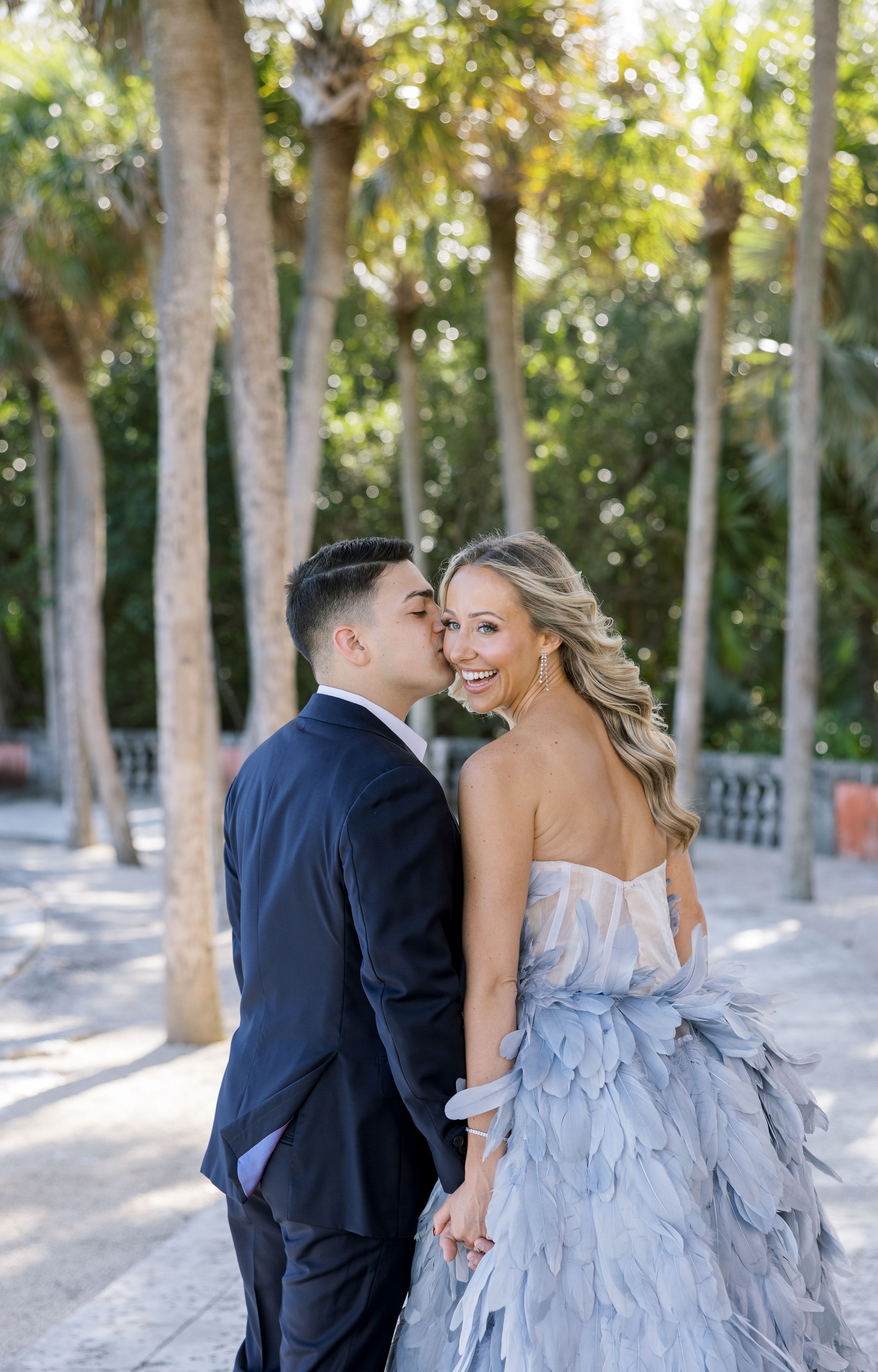 A romantic couple in formal attire poses among palm trees with a stunning blue layered gown.