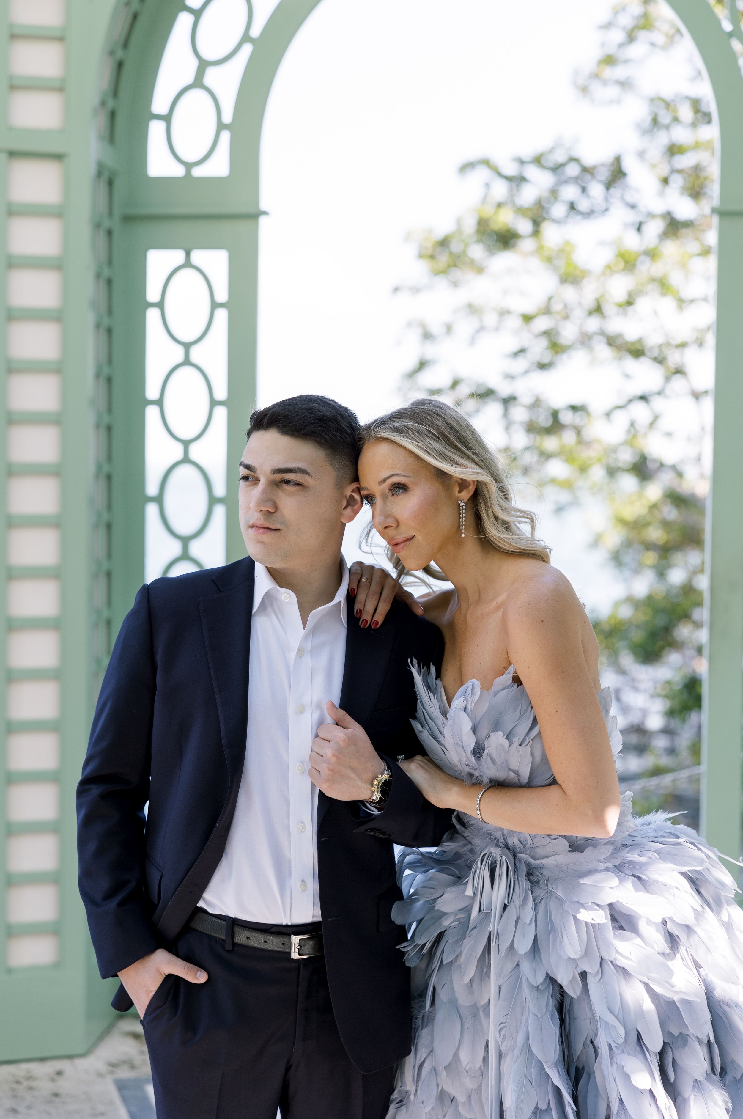 A stylish couple poses in formal attire beneath a mint green archway overlooking water.