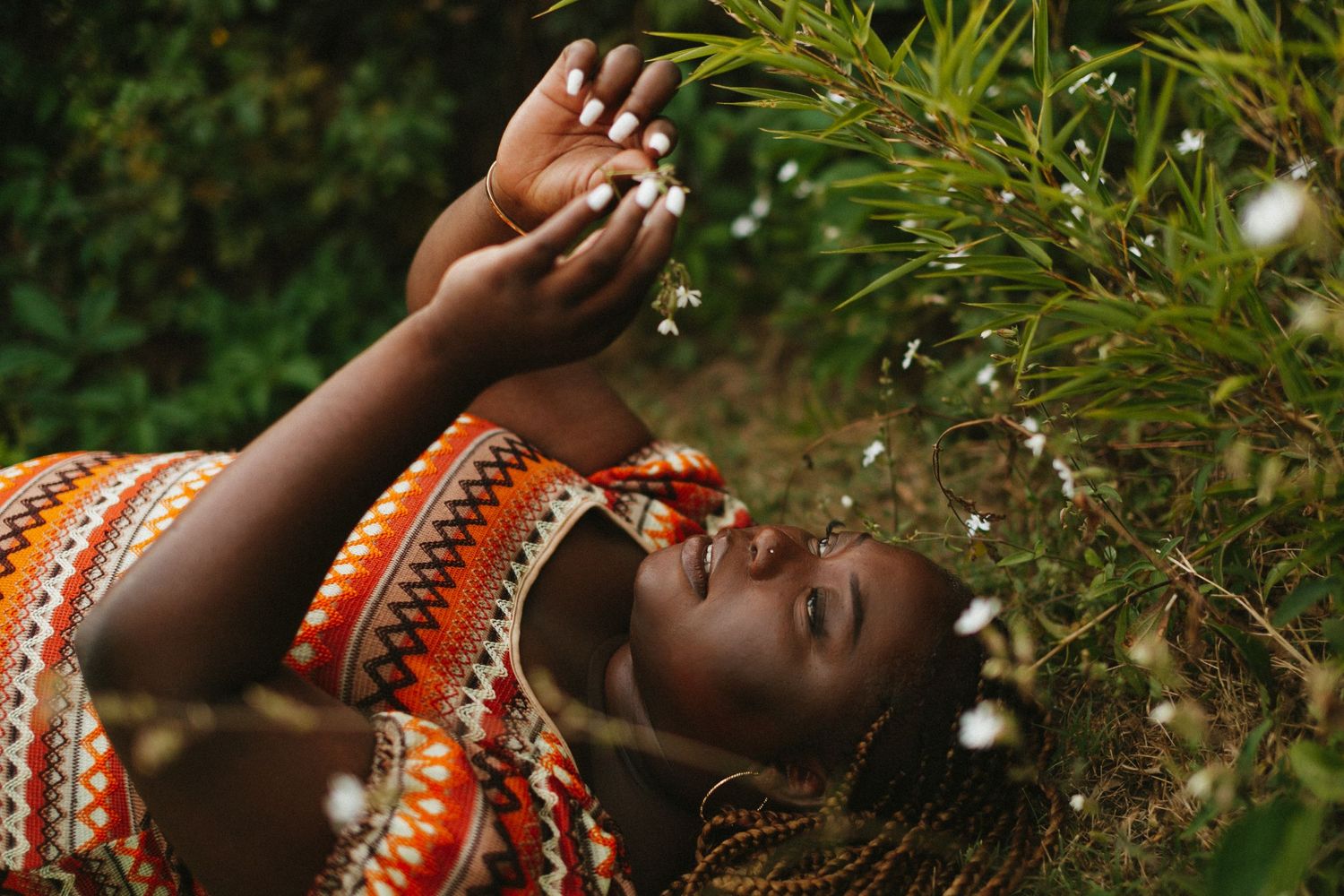 A person in an orange patterned top lies on grass reaching toward delicate white flowers in a dreamy nature scene.