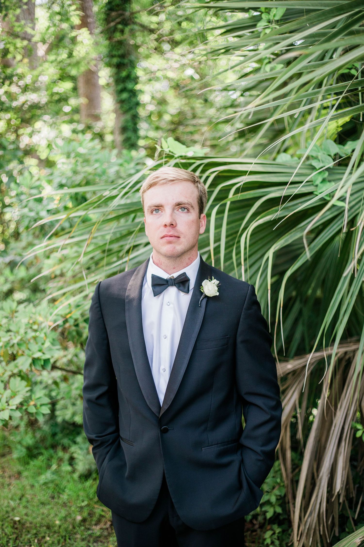 A groom in classic black tuxedo poses against tropical greenery backdrop.