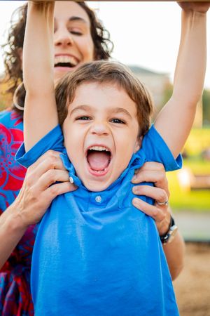 A curly-haired Mom laughs as she helps her young son with the monkey bars at a DC playground.