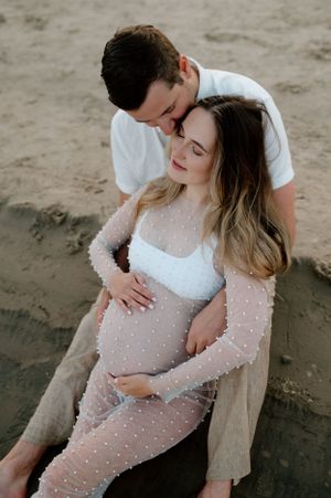 Tender maternity moment on beach with couple in white and sheer dotted dress.