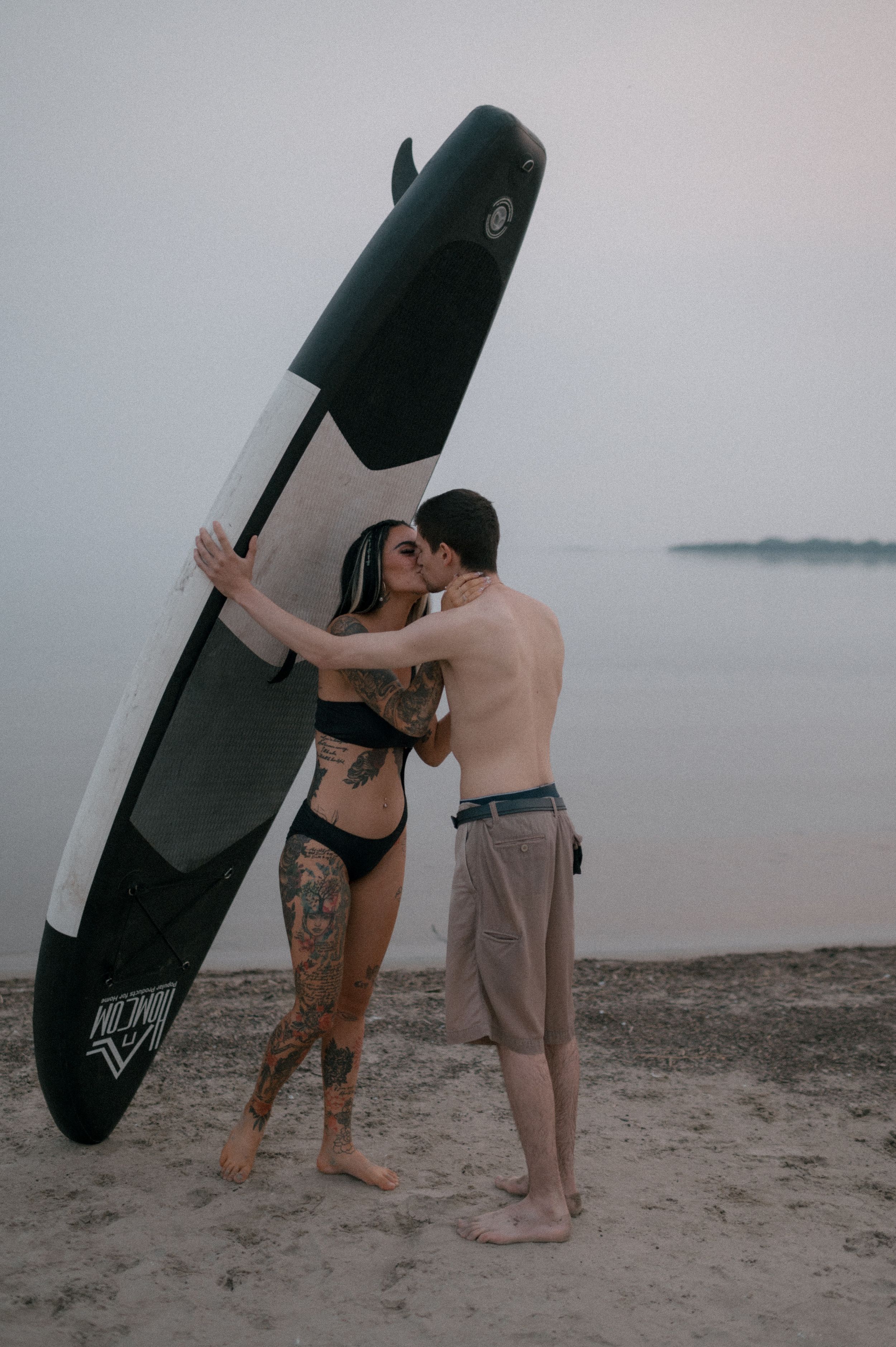 Two people on a beach with a surfboard at dusk.