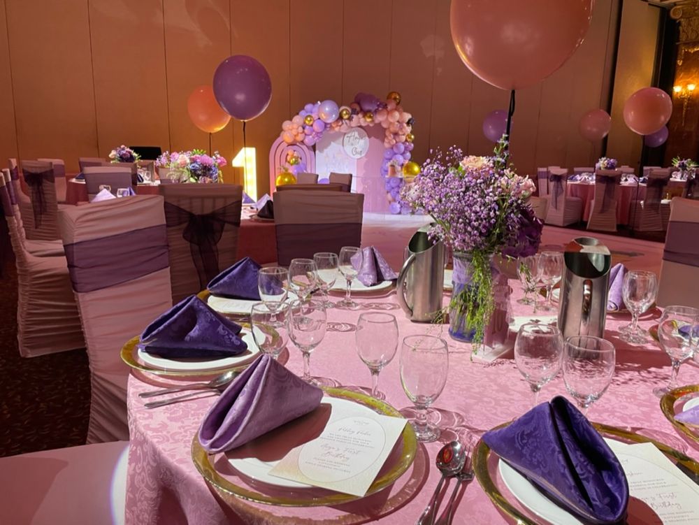 Guest table setup for a birthday party with pink and purple flowers and balloons, and cutlery