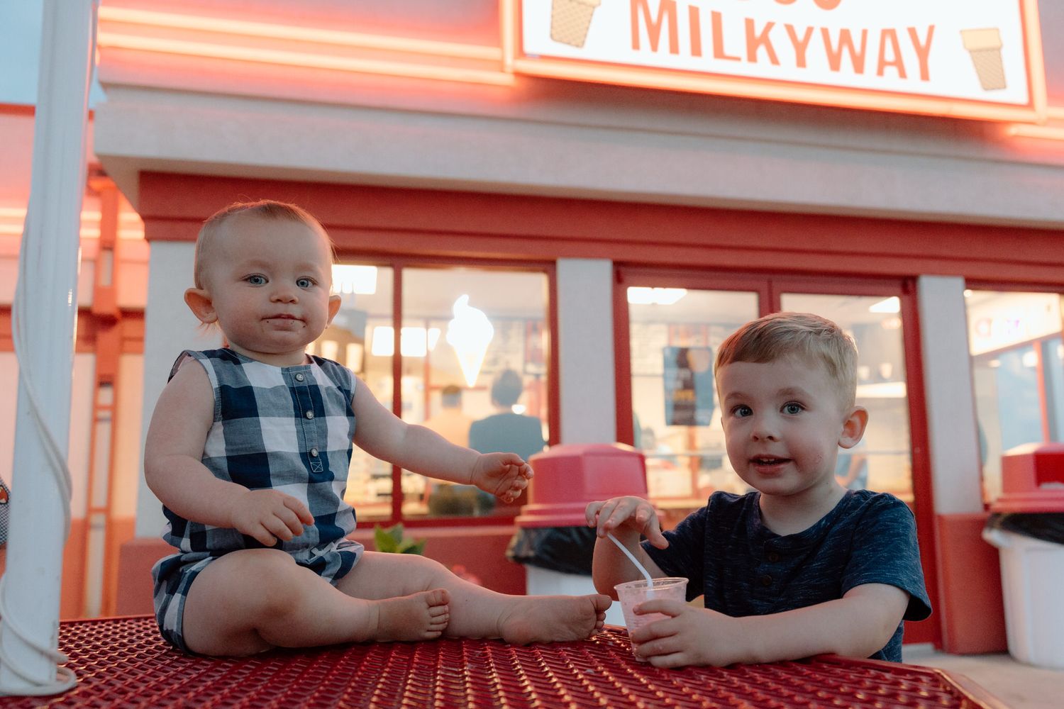 Two children sitting at red table outside Milkyway ice cream shop.