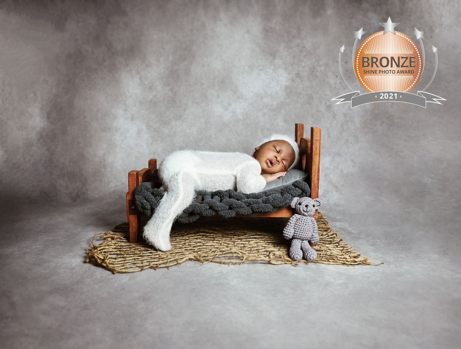 A sleeping toddler in white clothing lies on a small wooden bed with a plush toy on straw bedding.