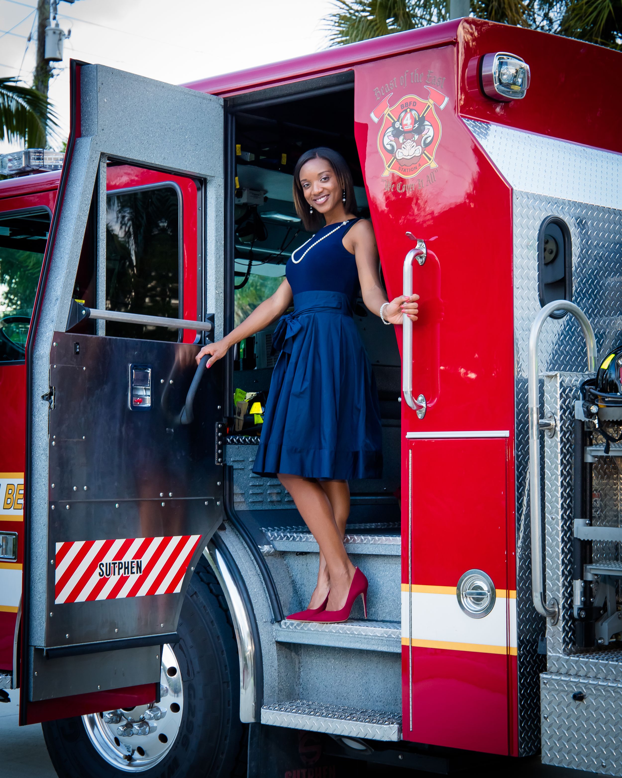 A person in a navy blue dress poses on the steps of a bright red fire truck.