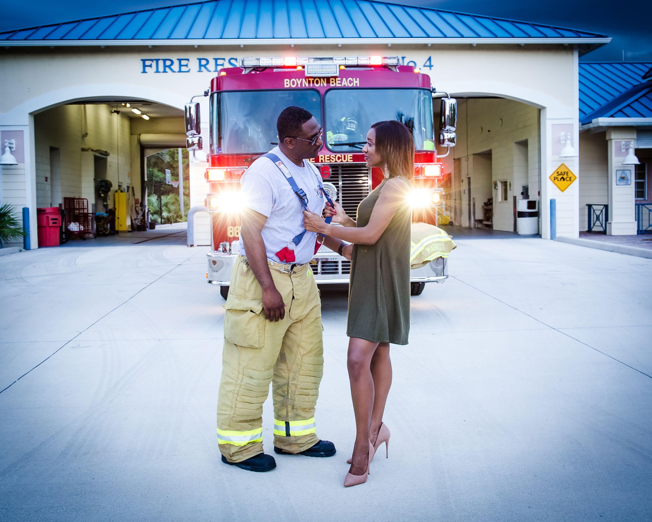 A firefighter and a person in a dress stand in front of a fire station with truck in background.