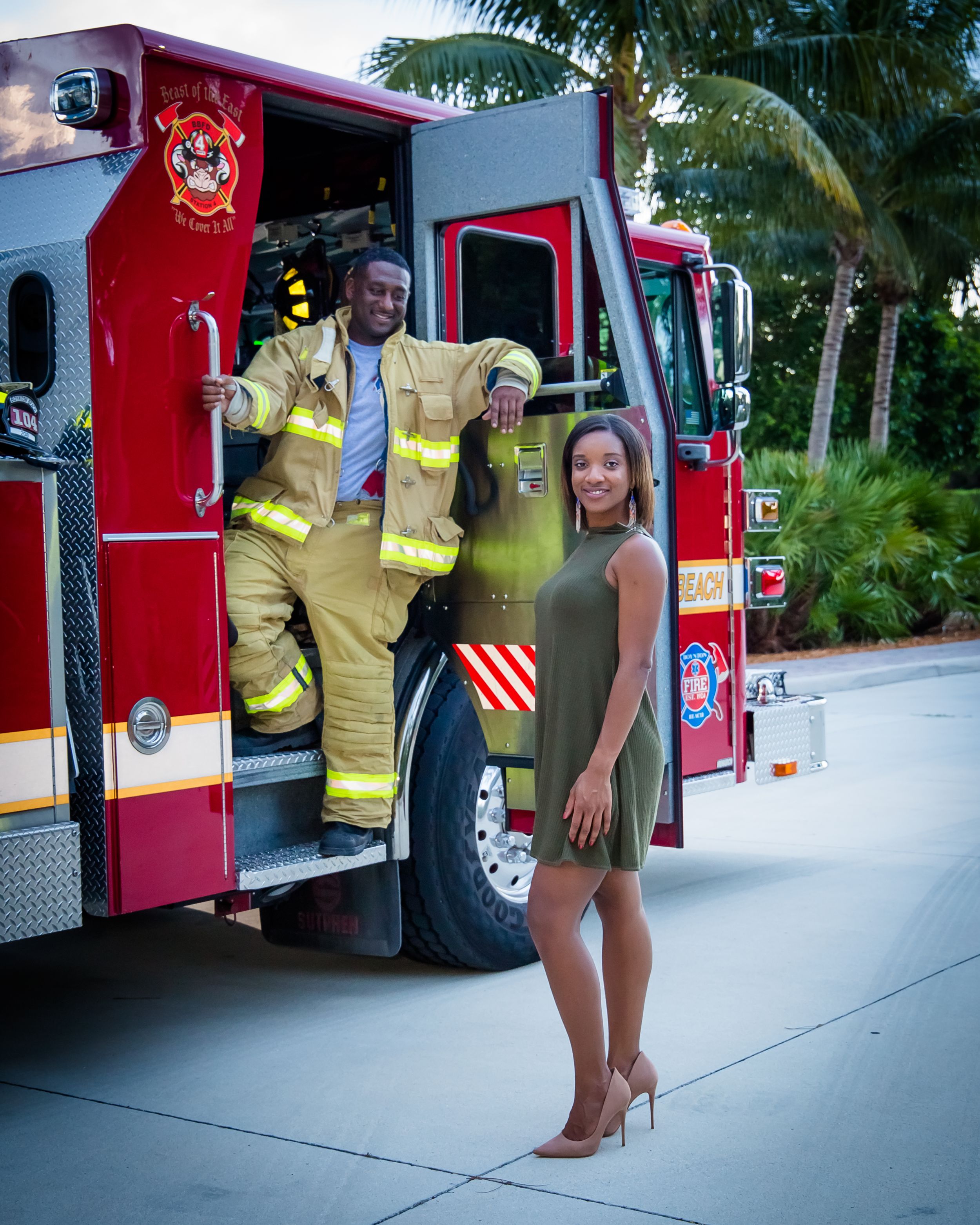 A firefighter in yellow gear and a person in a dress pose by a red fire truck.