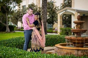 Two people share an intimate moment near an ornate water fountain surrounded by greenery.