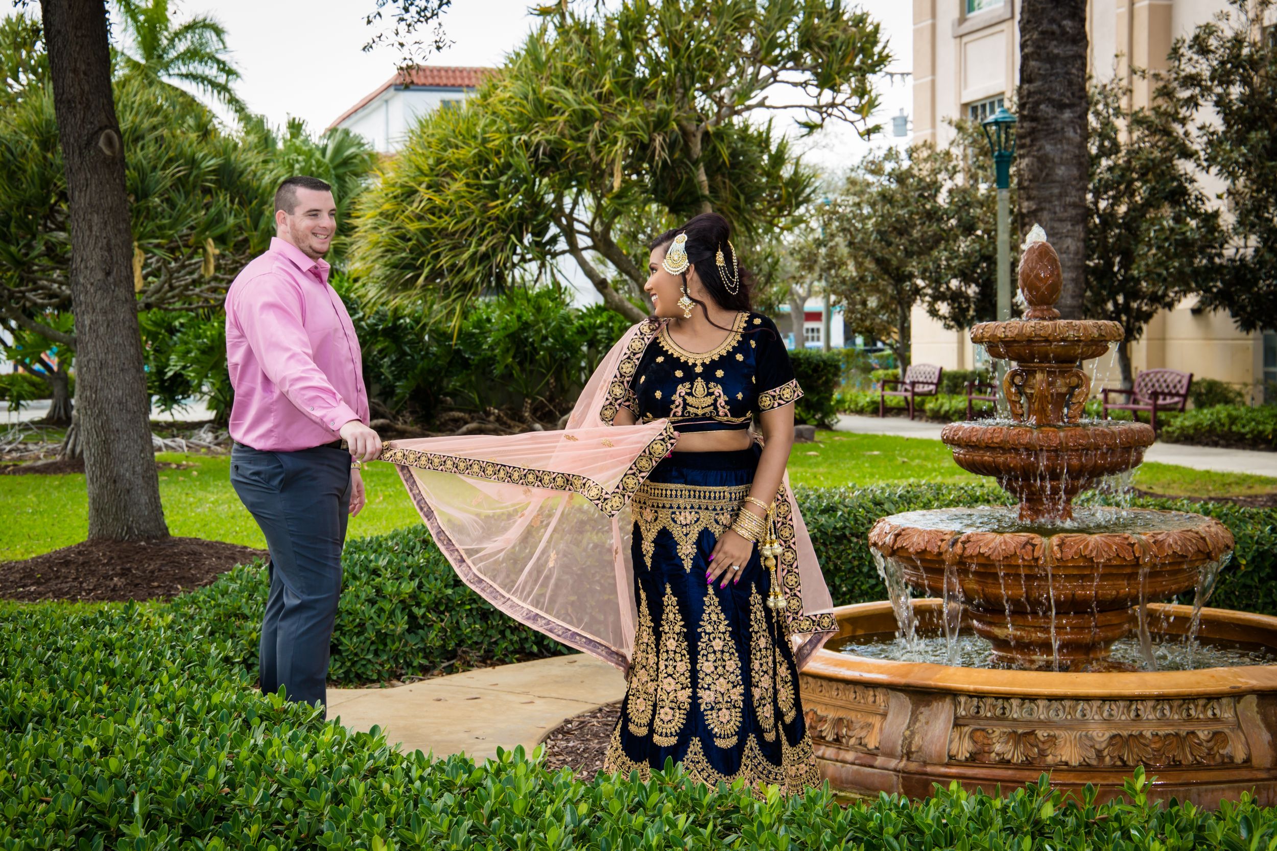 A romantic couple poses by a decorative stone fountain in a lush garden setting.