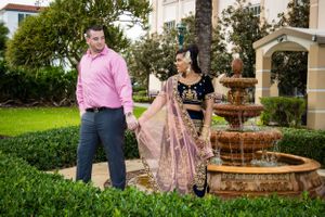 A couple in coordinated outfits poses together by a tiered fountain in a garden setting.