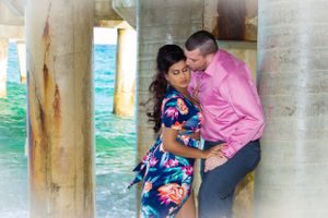 A couple in pink and floral outfits share an intimate moment near white pillars by the water.