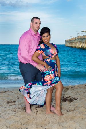 A couple sits together on a sandy beach with a pier and bright blue ocean in the background.