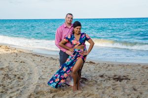 Two people pose playfully on a beach with turquoise ocean waters in the background.