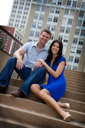 A couple sits casually on concrete steps with an urban backdrop of tall buildings.
