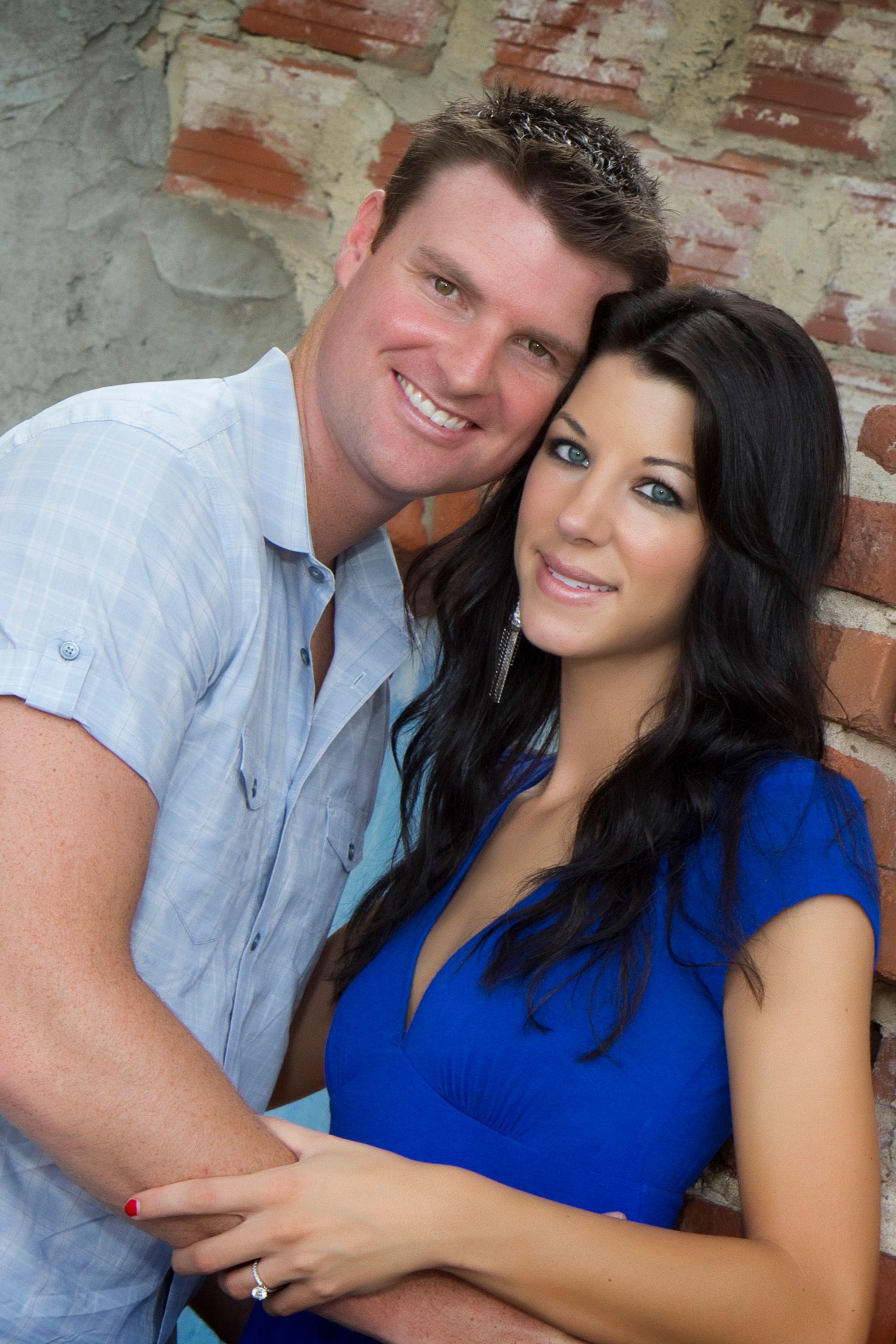 A close-up portrait of a couple in casual attire against a brick wall background.