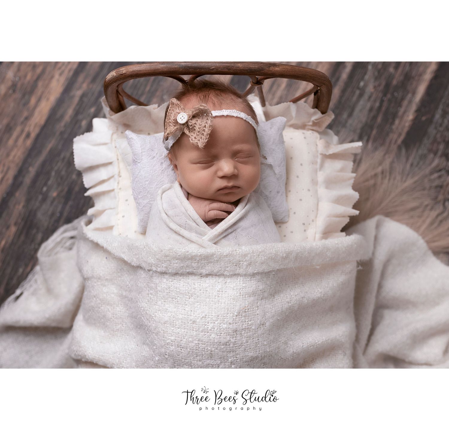 Sleeping newborn wrapped in white textured fabric with pink bow in rustic setting.