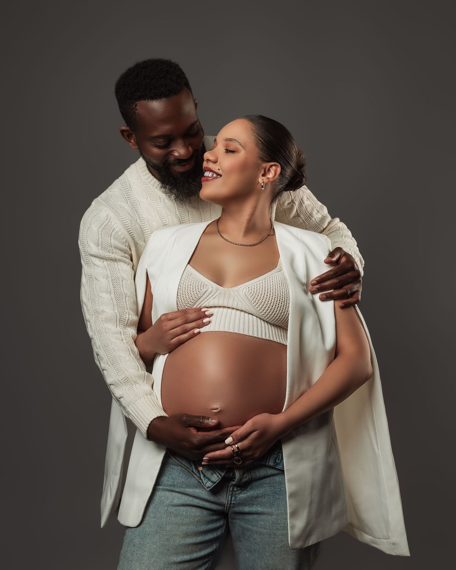 Intimate maternity photography of couple in white knitwear against gray studio background.