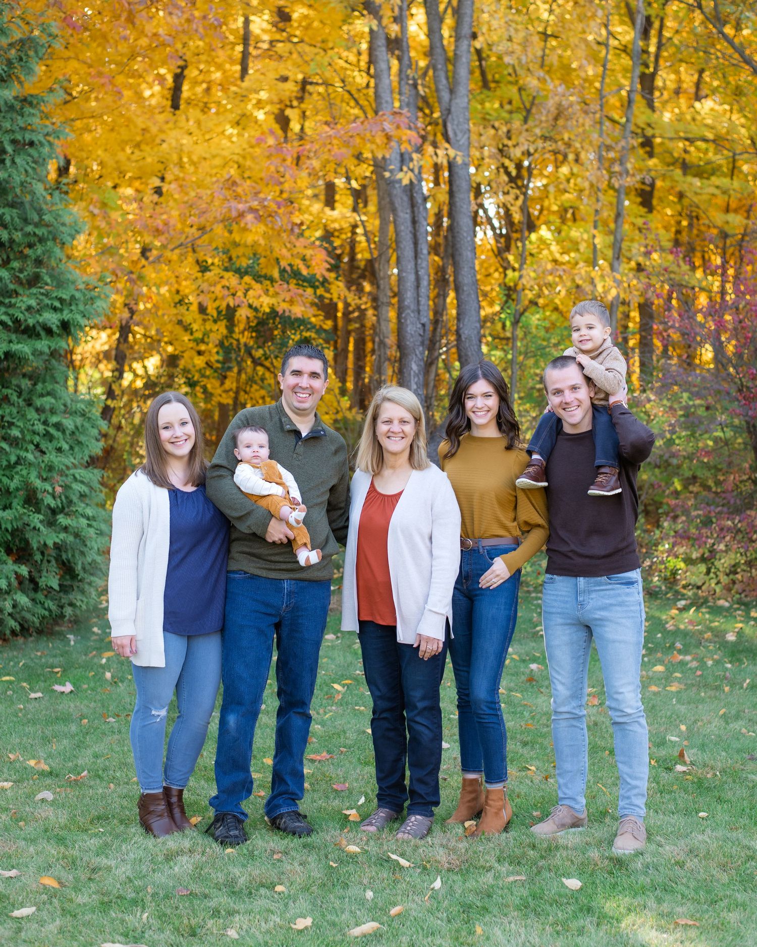 Extended family members gather for a portrait outdoors against a backdrop of golden fall trees.