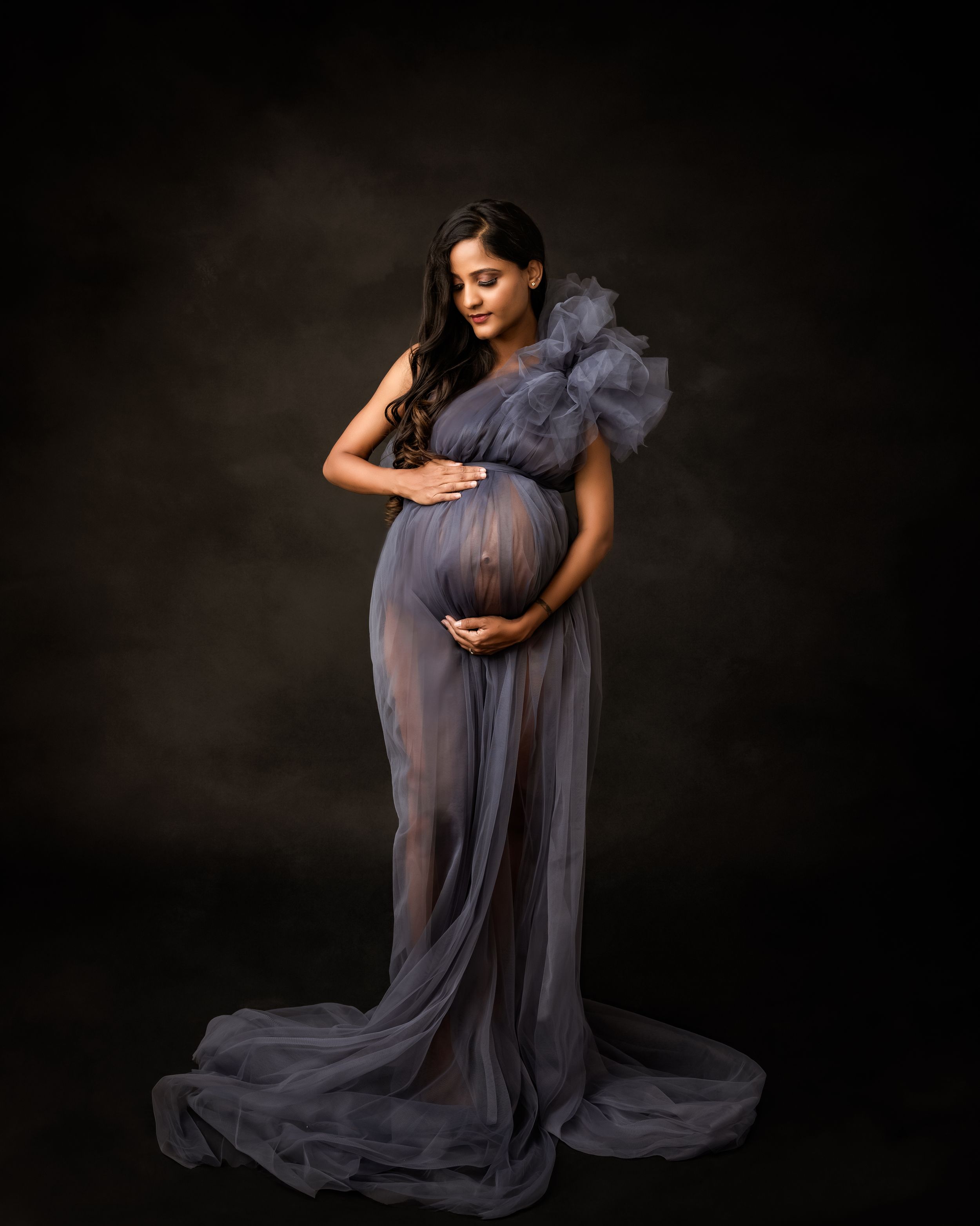Maternity portrait in a flowing slate blue gown with ruffled shoulders against dark backdrop.