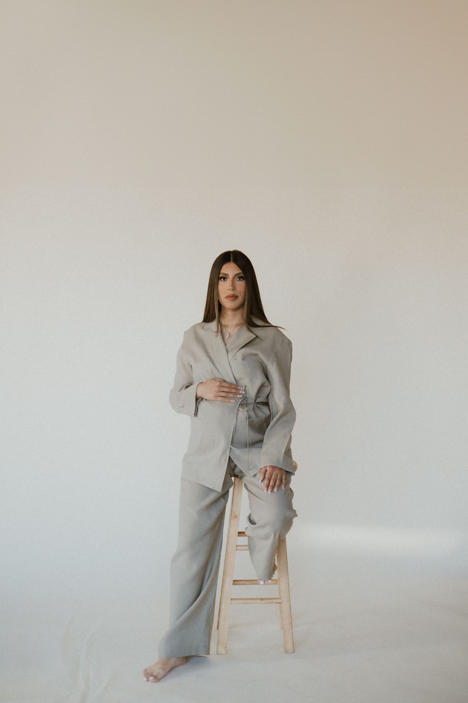 A minimalist portrait of someone seated on a wooden stool wearing an oversized gray suit.