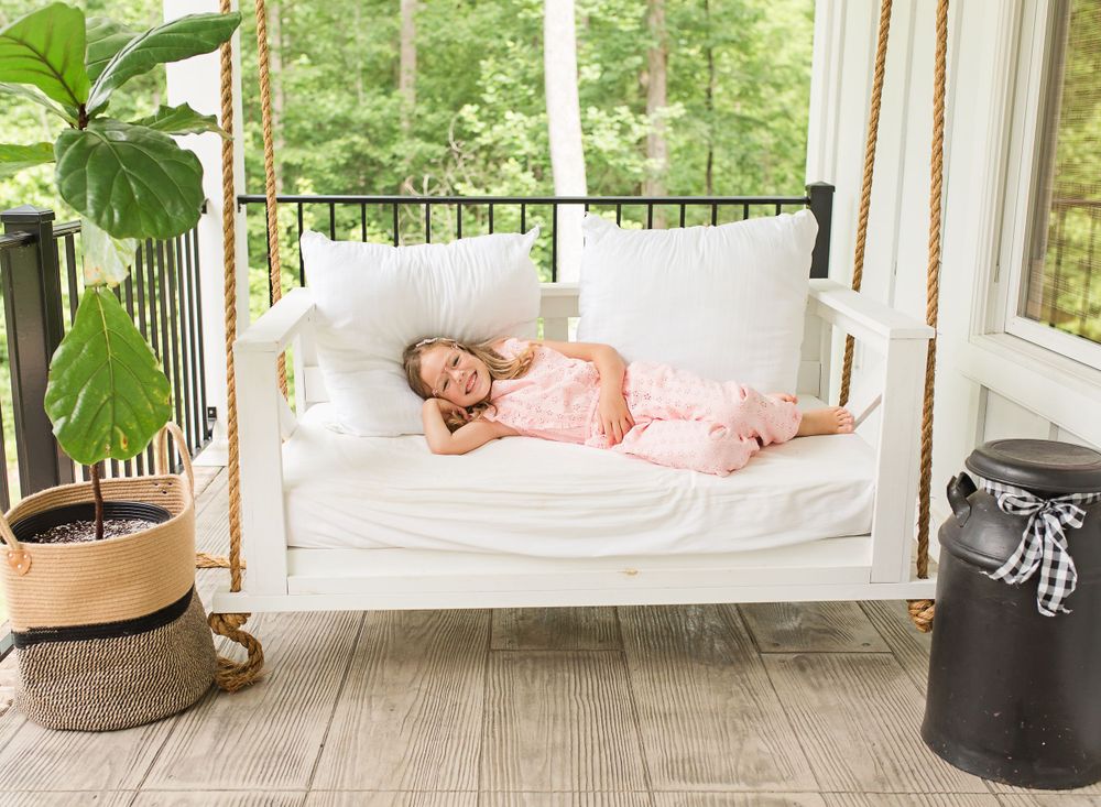 A child relaxes on a white hanging bed on a sunlit porch with indoor plants.