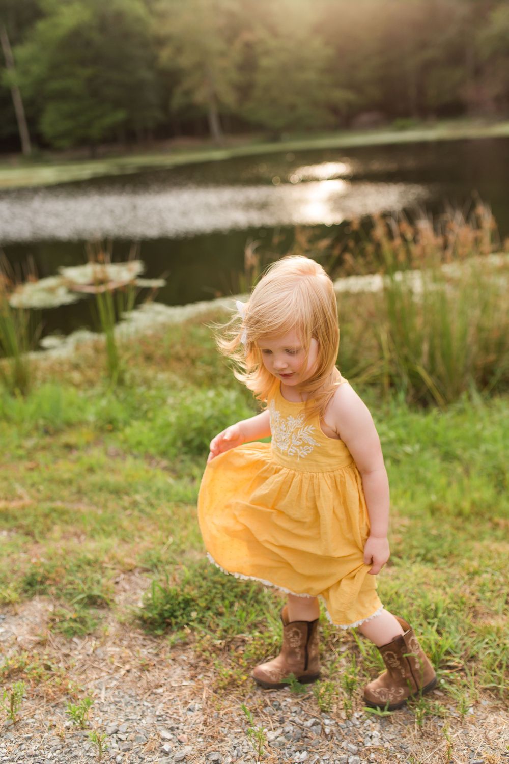 Young child in yellow dress twirling by a pond at sunset wearing brown boots.
