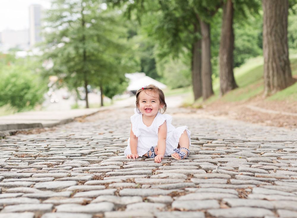 A joyful child in a white dress sitting on a cobblestone path surrounded by green trees.