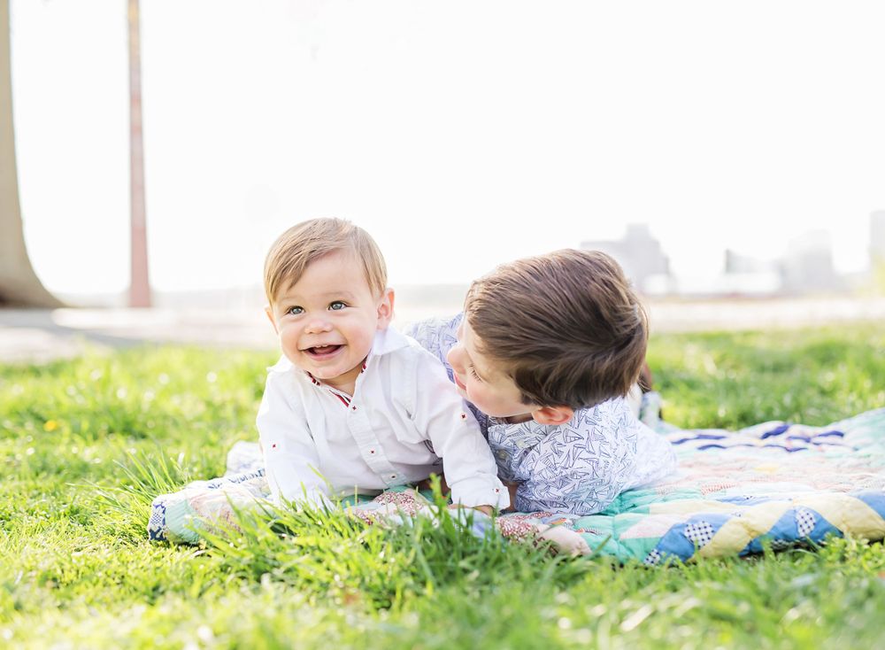 Two children play together on a blanket in the grass on a sunny day.