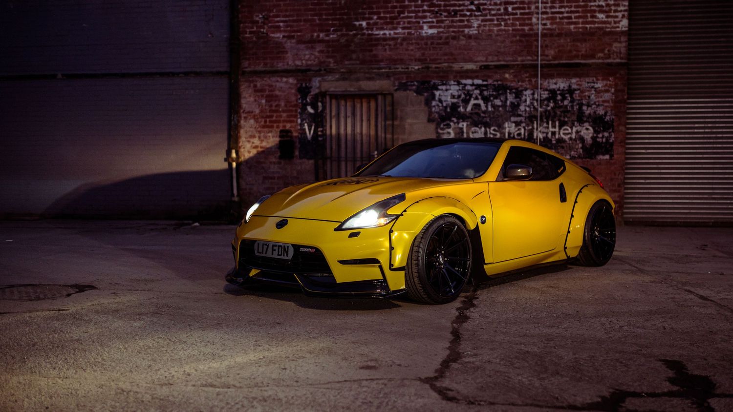 A bright yellow modified Nissan sports car parked in front of a dark brick wall at night.