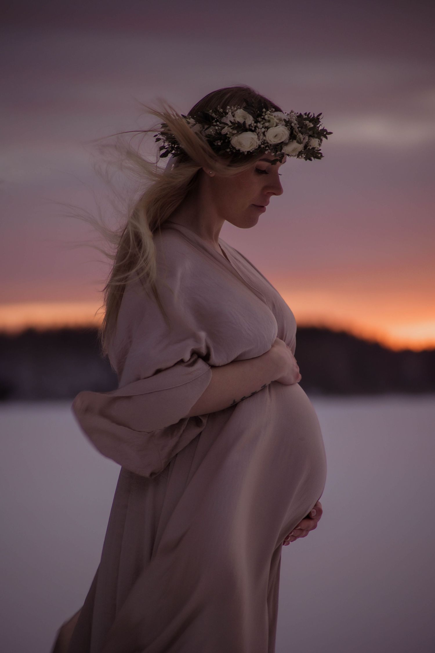 Pregnant woman in flowing blush gown and white floral crown stands in snowy landscape at sunset.