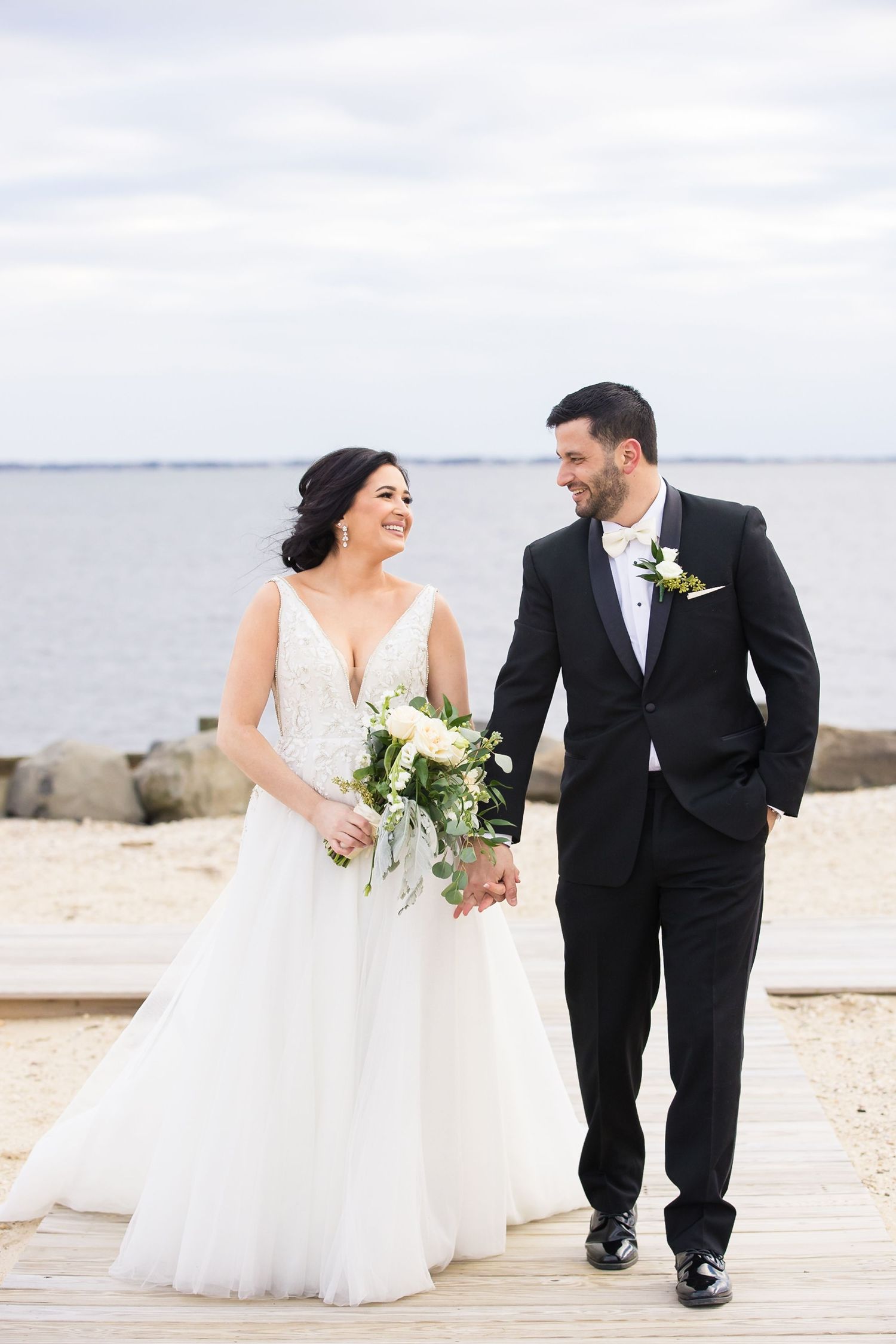 A couple in wedding attire walks together on a waterfront boardwalk holding a white floral bouquet.