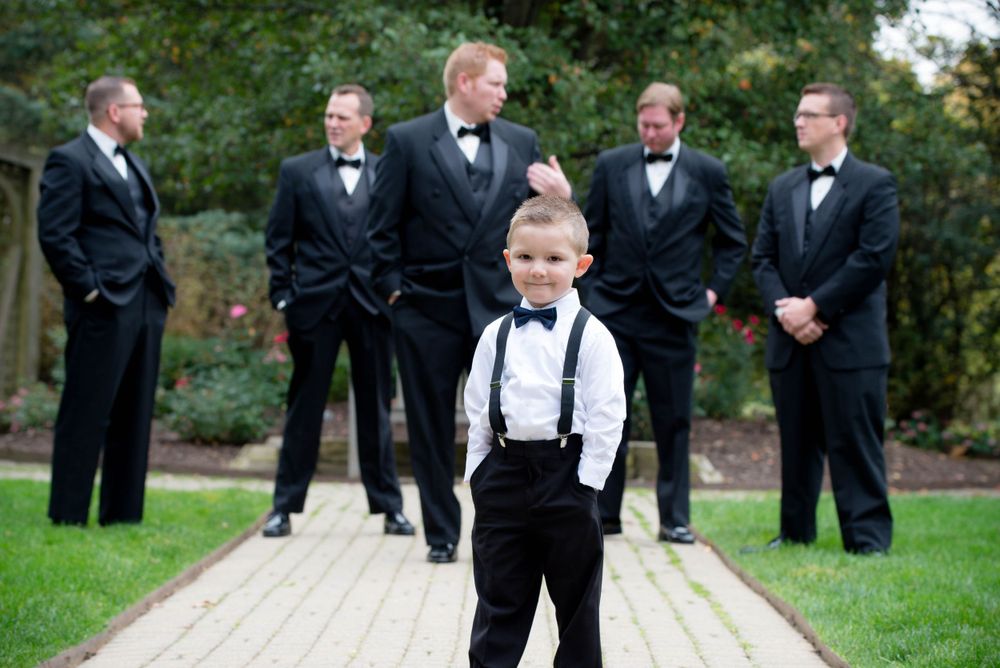 Ring bearer in suspenders poses confidently with groomsmen in background.