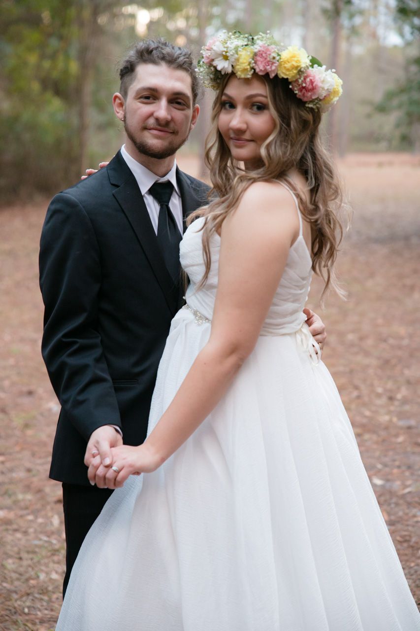 Newlyweds pose in a forest setting with bride wearing floral crown and white gown.