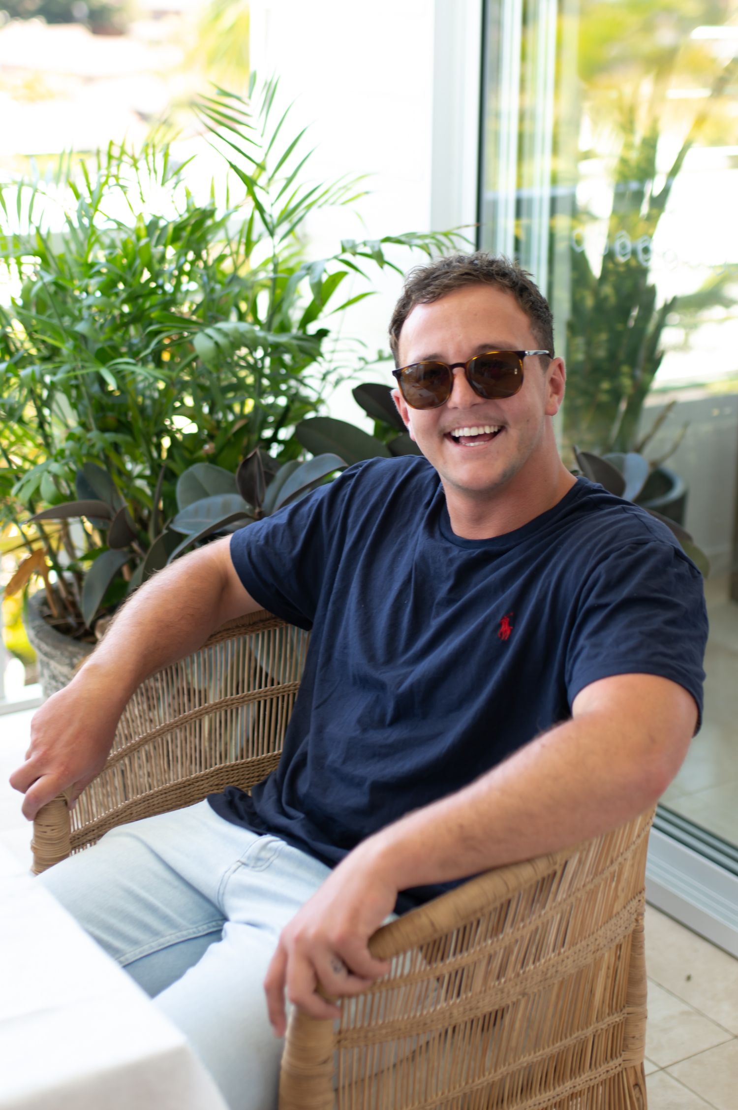 Someone relaxing in a wicker chair on a sunny patio wearing sunglasses and a navy polo shirt.