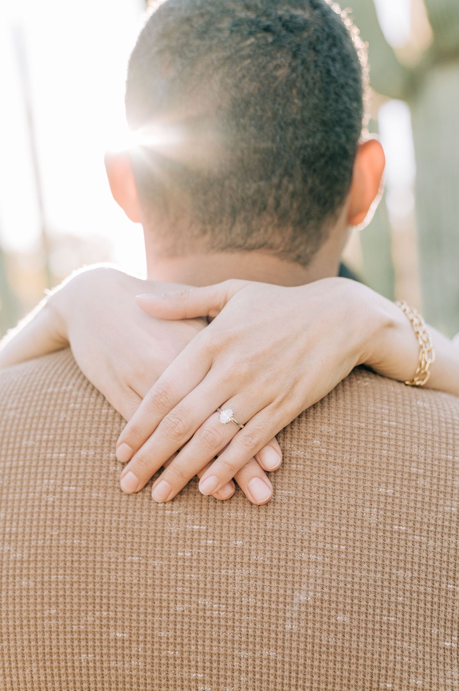Couple embracing outdoors, woman's hand resting on man's shoulder displaying a diamond engagement ring, golden bracelet, sunlight.