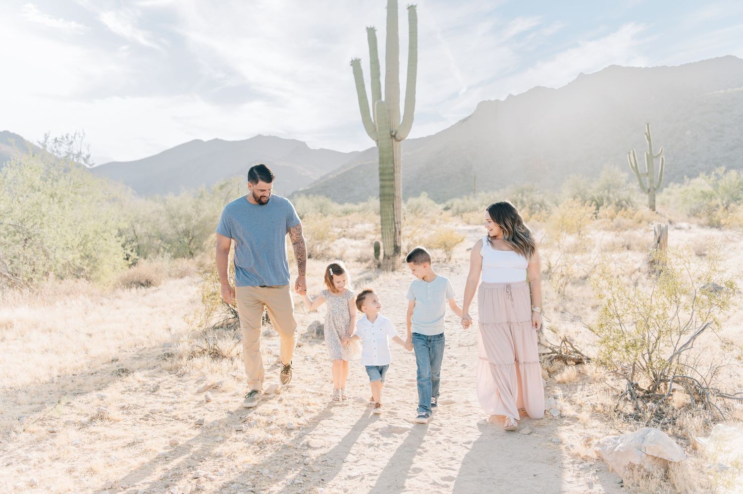Family of five walking hand-in-hand through Arizona desert with saguaro cactus and mountains in background.