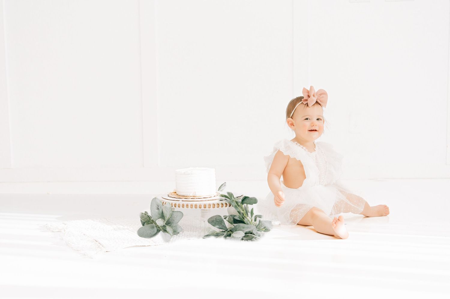 Baby girl in white dress with pink bow sits on white bedding beside eucalyptus and teacup tray.