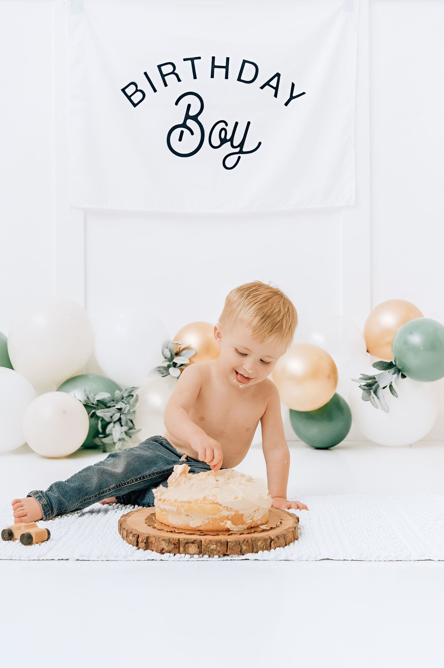 Toddler boy smashing his first birthday cake at a cake smash photoshoot with balloons and Birthday Boy banner.