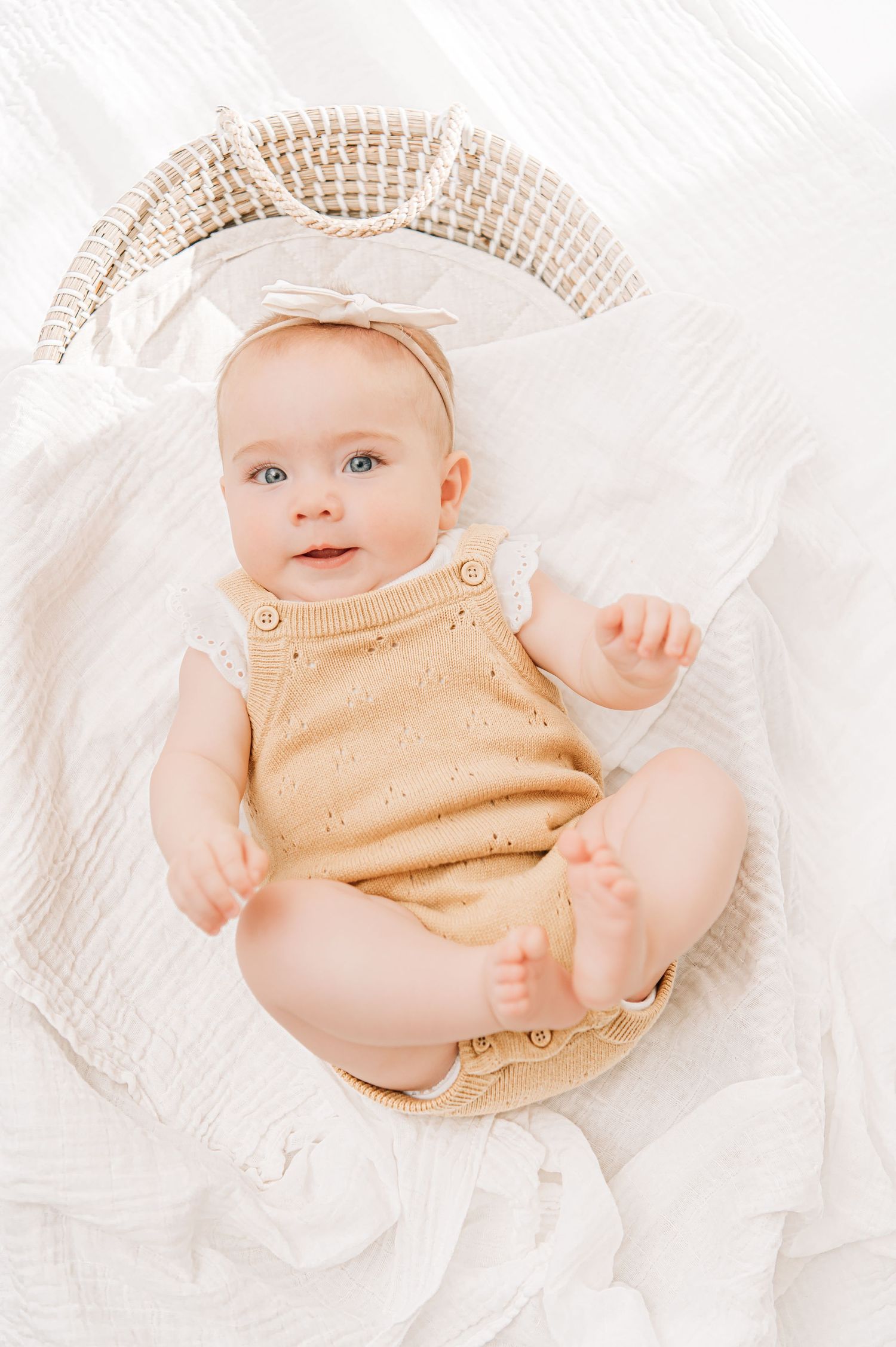 Smiling baby girl in yellow knit romper and white bow headband lying in a wicker basket on white blankets.