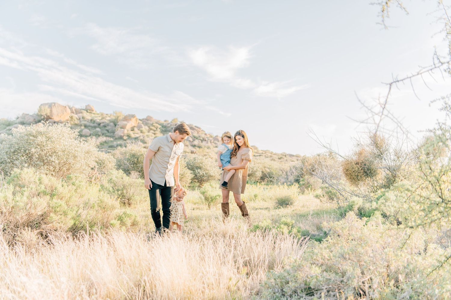 Family of three walking through a sunlit desert landscape with rocky hills and golden grass in the background.