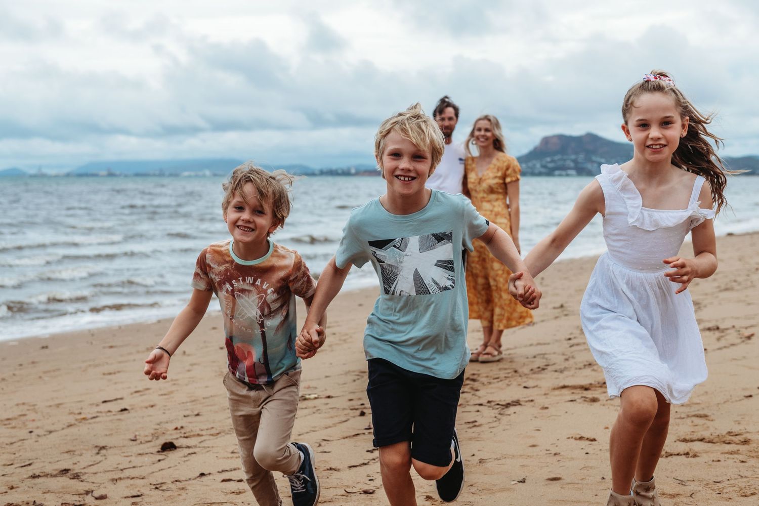 Children running joyfully along a sandy beach with mountains in the background.